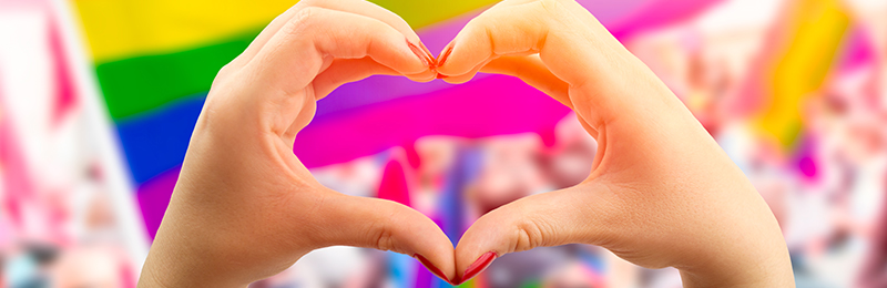 A background of a gay pride flag framed by a person's hands brought together in the shape of a heart in the foreground 