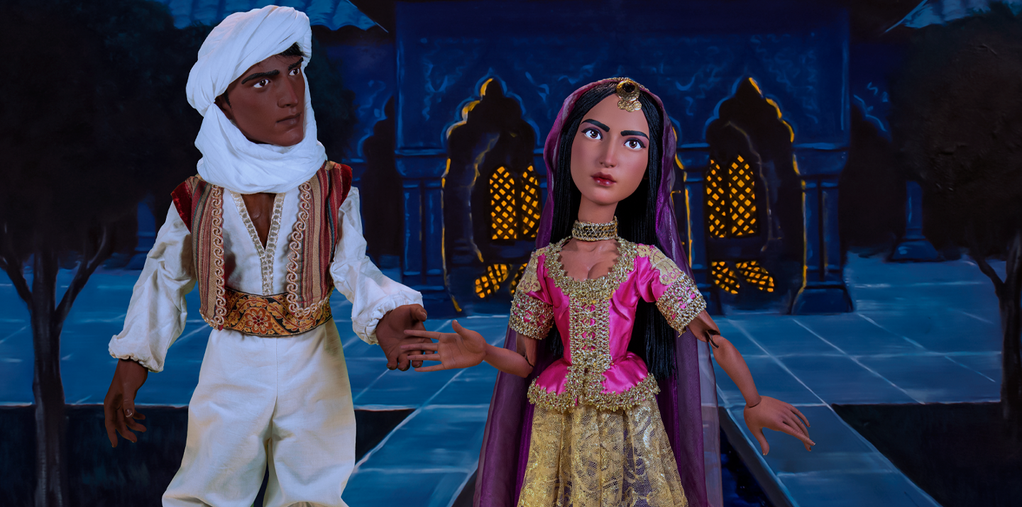 A male puppet wearing all white with a red patterned vest and a white head wrap holds the hand of a female puppet wearing pink and gold with a purple veil.