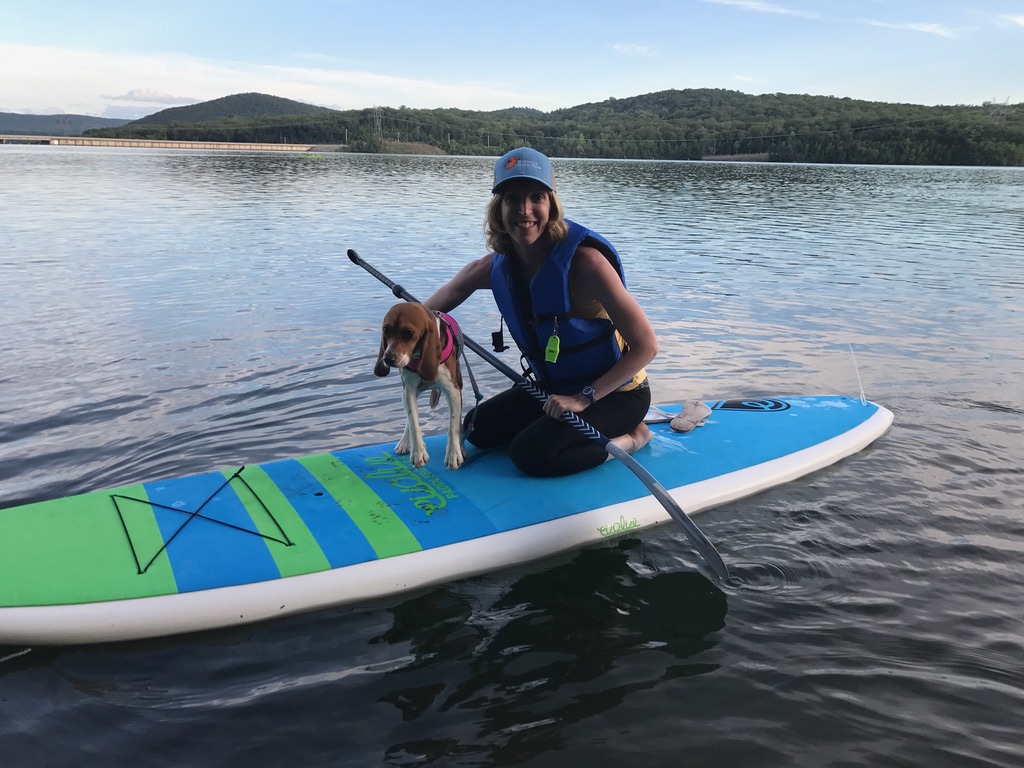 Woman paddleboards with a beagle on a large lake with mountains in the background