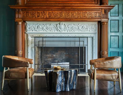 Stone fireplace with wooden mantel, flanked by two leather club chairs, in great room of mansion